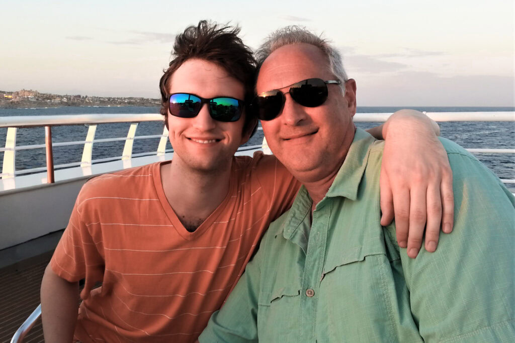 Two men wearing sunglasses smile and pose together on a boat deck at sunset. One man has his arm around the other. The ocean and coastline are visible in the background.