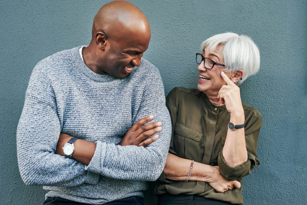 A smiling man and an older woman with glasses stand against a blue wall, laughing and looking at each other. The man wears a gray sweater, and the woman wears an olive green shirt.
