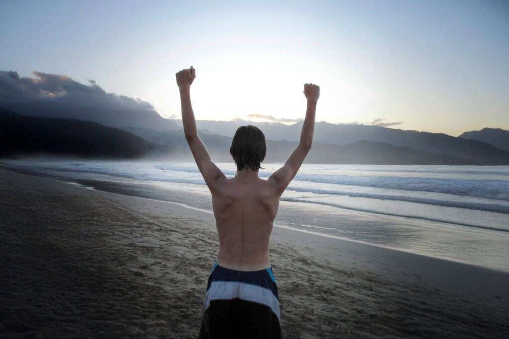 A person standing on a beach at sunrise or sunset with arms raised in triumph, facing the ocean and distant mountains.