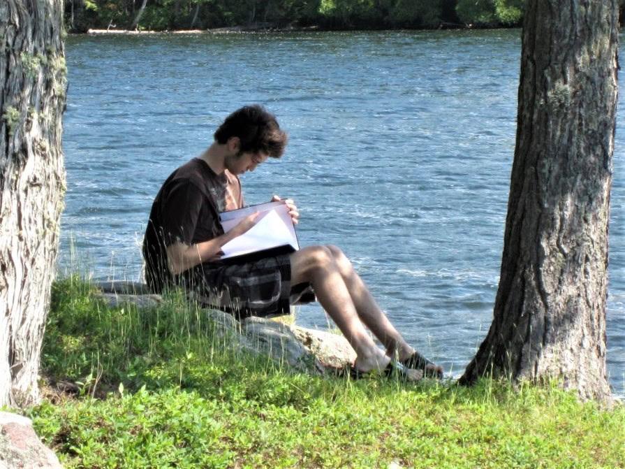 A person sits on a rock between two trees near the edge of a lake, writing or drawing in a notebook with sunlight filtering through the trees and water visible in the background.