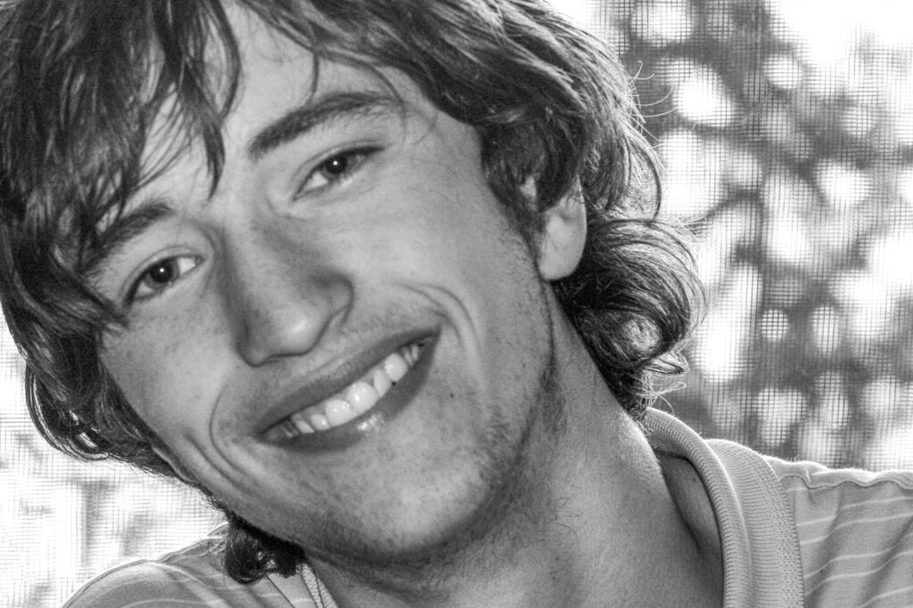A young man with wavy hair smiles broadly at the camera. He is wearing a light-colored, collared shirt. The background is blurred, showing some trees and light coming through a window. The image is in black and white.