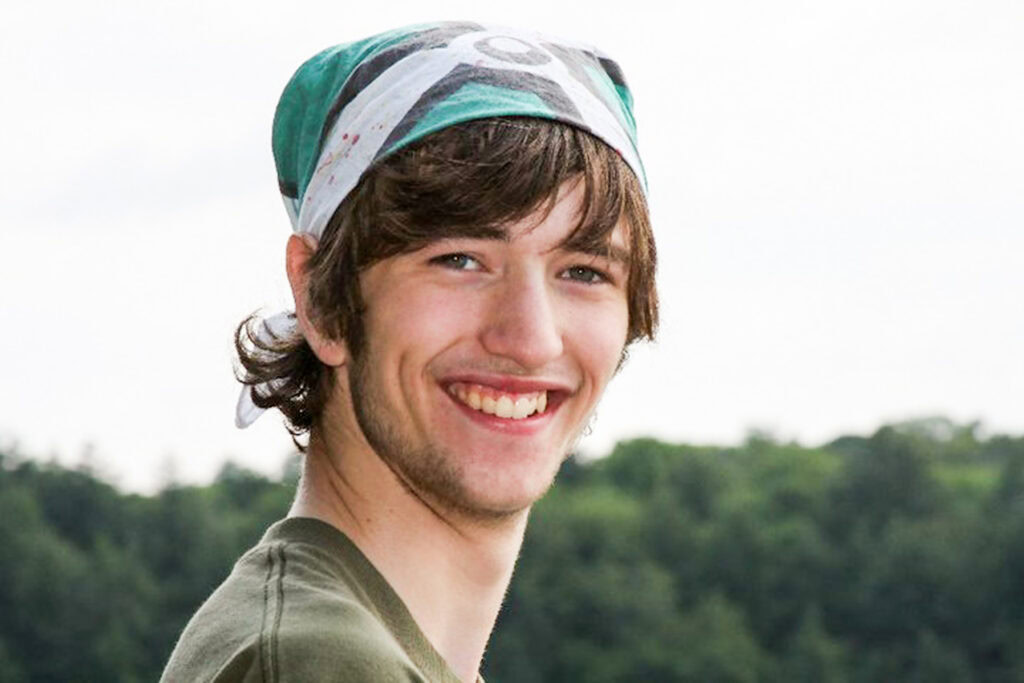 A young man with long brown hair and a beard smiles at the camera, wearing a green shirt and a patterned bandana on his head. There are trees and a cloudy sky in the background.