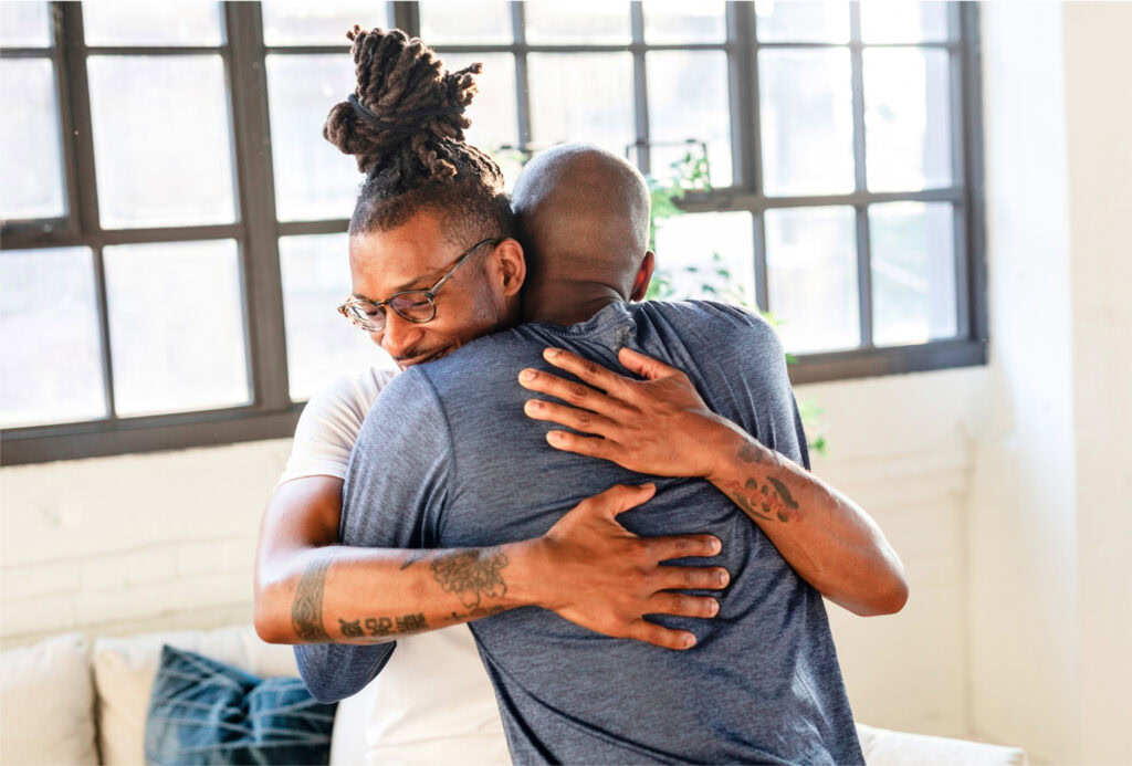 Two men are warmly hugging each other in a brightly lit room with large windows. One man wears glasses and a white shirt, while the other wears a blue shirt. Both appear happy and comfortable.