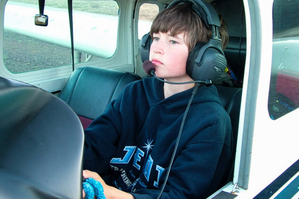 A young boy wearing a headset sits in the cockpit of a small airplane, looking forward and holding the controls while dressed in a dark hoodie.