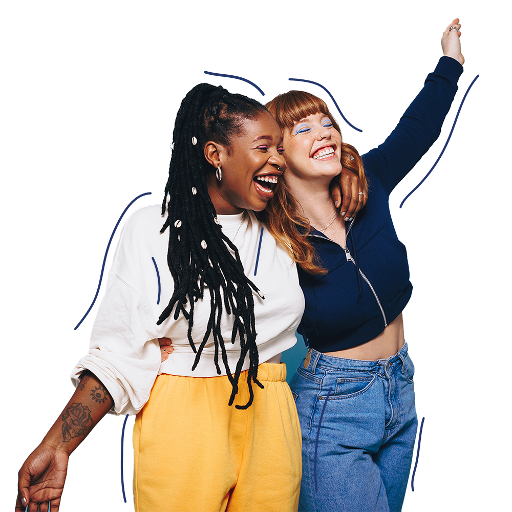 Two women smiling and laughing together, one with long dark braids and yellow pants, the other with red hair and a blue top, standing close with arms around each other against a white background.